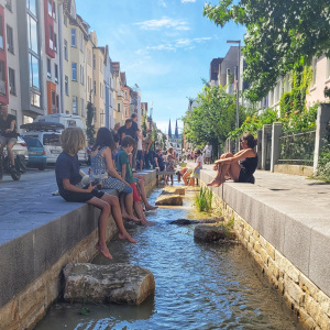 Kinder und Erwachsene sitzen am Lutterkanal in der Ravensberger Straße und lassen ihre Füße ins Wasser baumeln.