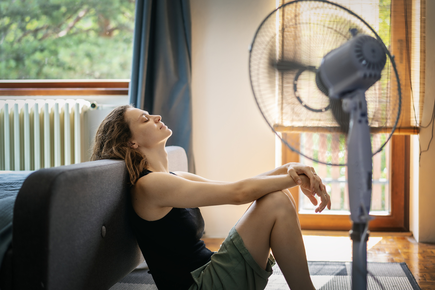 Junge Frau, die unter Hitze leidet, sitzt zu Hause auf dem Boden vor einem Ventilator.