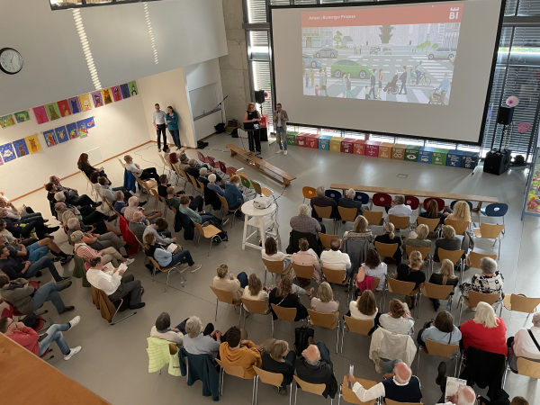 Rund 80 Personen sitzen auf Stühlen und schauen auf eine große Leinwand. Davor stehen eine Moderatorin und ein Referent.