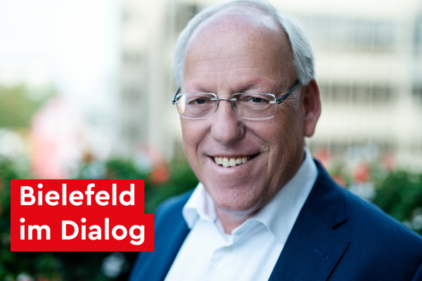 Foto vom Bielefelder Bürgermeister Pit Clausen (2024). Er lächelt in die Kamera, trägt eine rahmenlose Brille, ein weißes Hemd und ein blaues Sakko. Er steht draußen auf einem Balkon. Neben seinem Gesicht der Text auf rotem Hintergrund: "Bielefeld im Dialog"